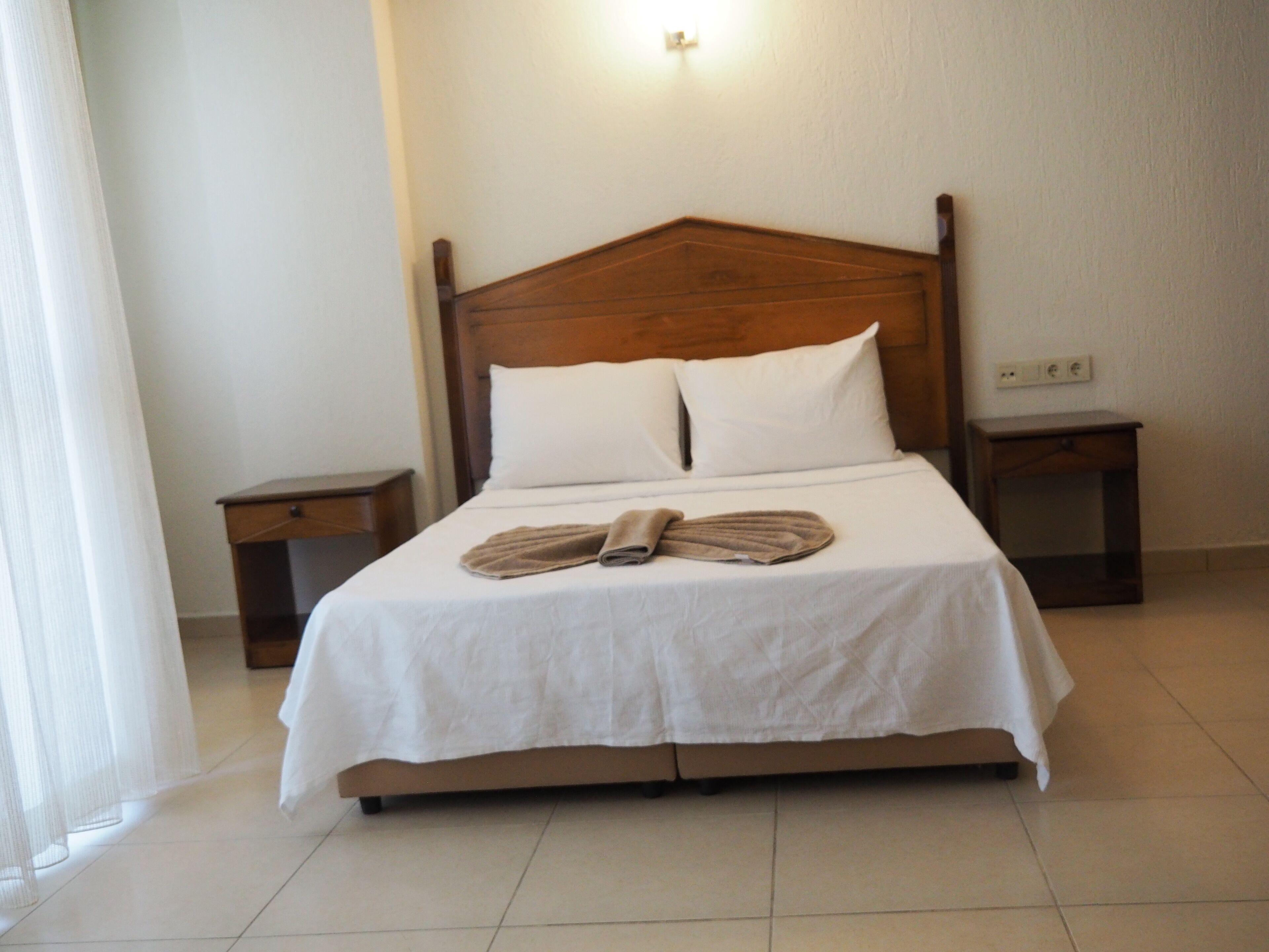 A neatly made bed with a wooden headboard, white bedding, and a folded towel on the bed. There are two bedside tables, one on each side of the bed.