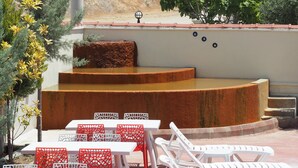 Una terraza al aire libre con mesas blancas y sillas rojas, un área de asientos escalonada y un fondo de árboles y un edificio.