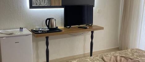 A hotel room with a bed, a desk with a kettle, a small fridge, a flat-screen TV, and a mirror.
