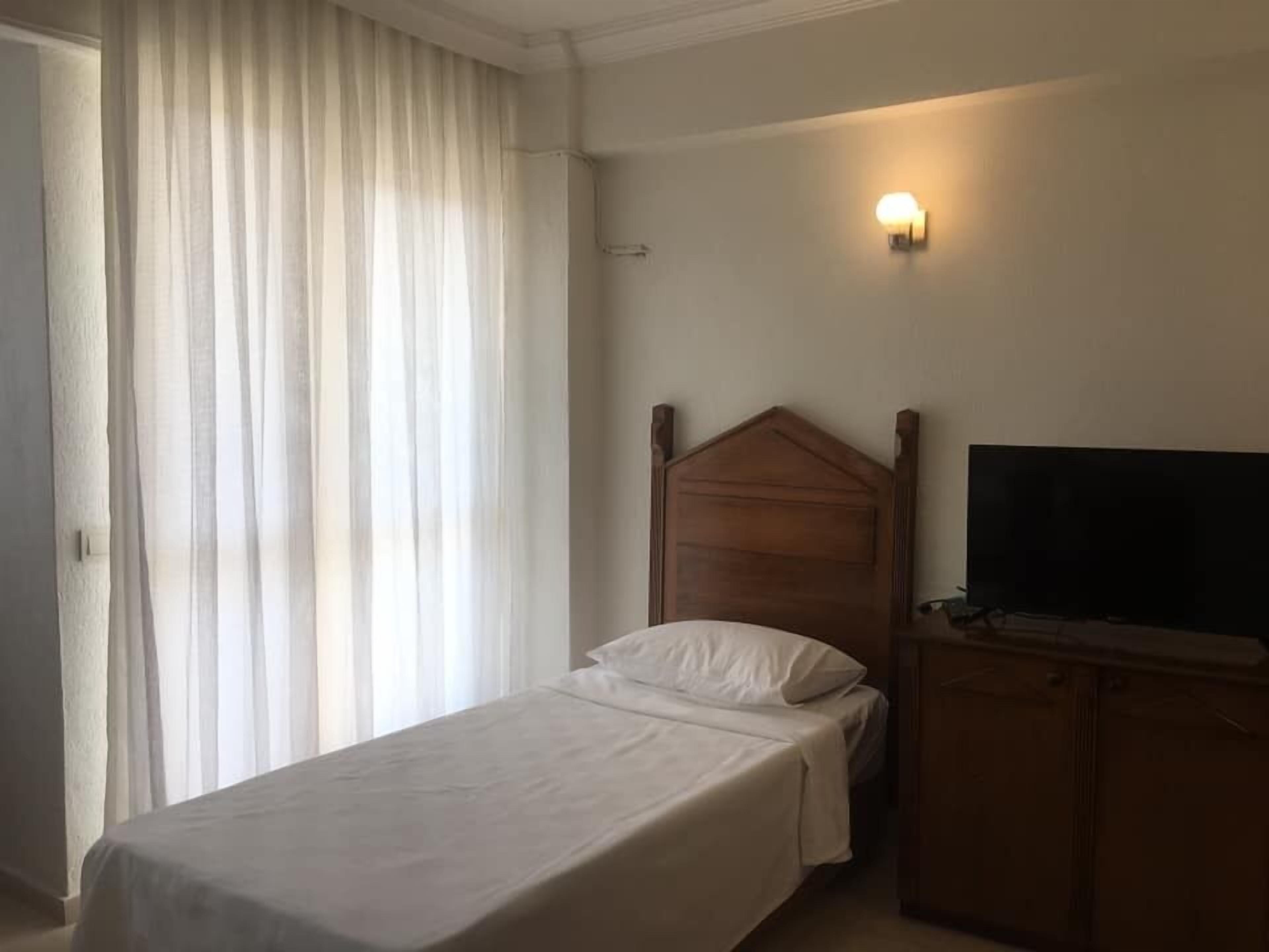 Een enkel bed met een houten hoofdbord, een televisie op een houten dressoir en een wandlamp.