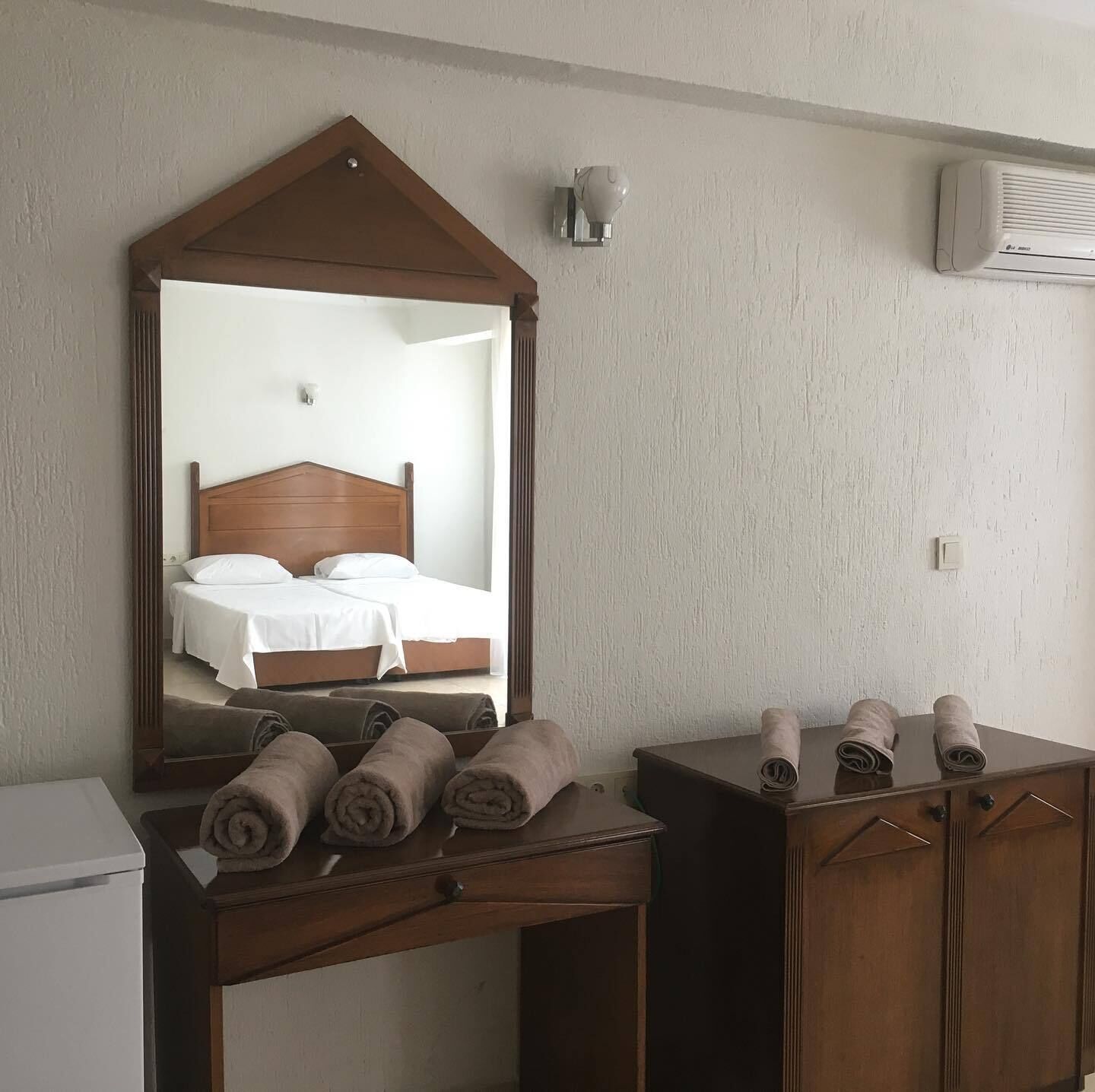 Quarto de hotel com cama de cabeceira de madeira, um espelho, uma escrivaninha com toalhas dobradas e um aparelho de ar condicionado na parede.