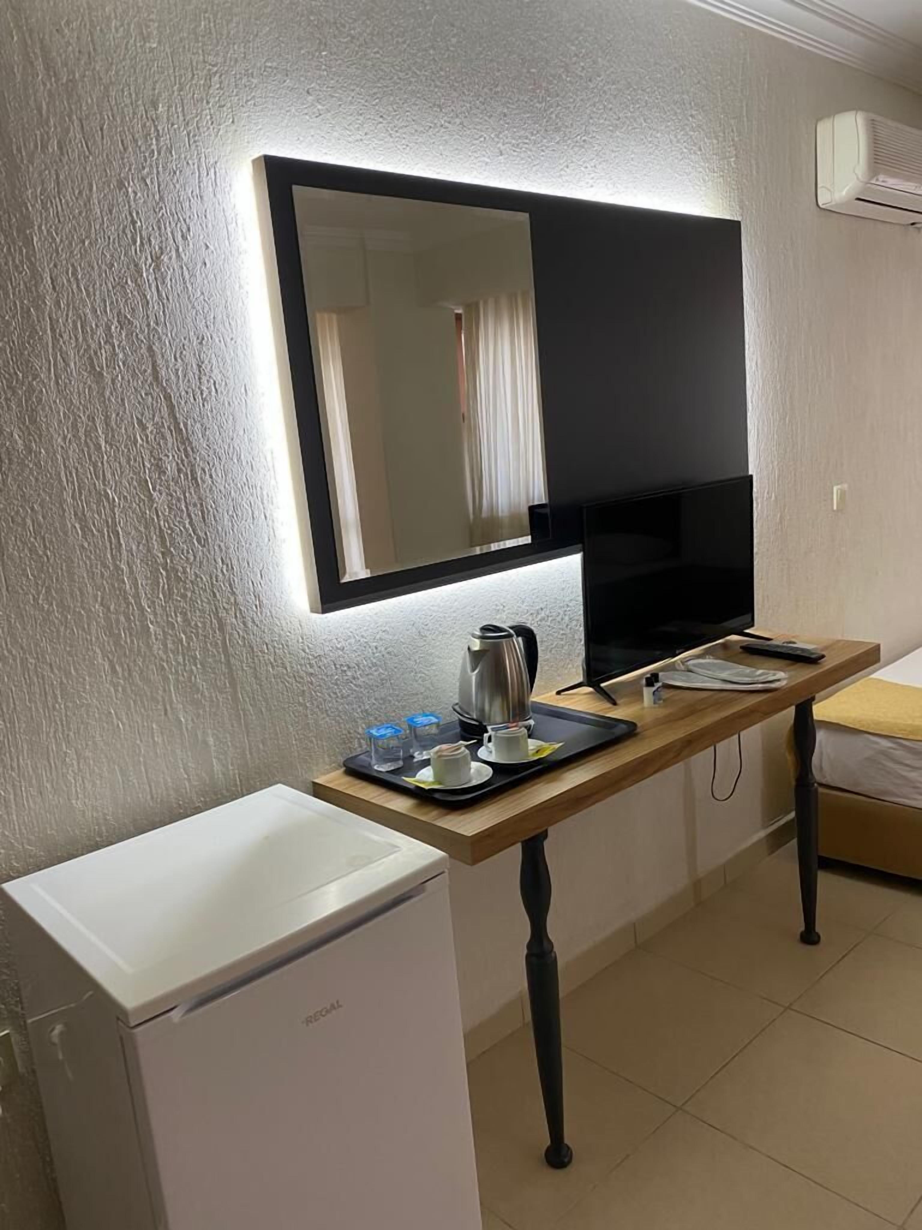 A compact hotel room with a bed, a small kitchenette, a flat-screen TV mounted on the wall, and a bedside table with a tray of breakfast items.