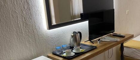 A compact hotel room with a bed, a small kitchenette, a flat-screen TV mounted on the wall, and a bedside table with a tray of breakfast items.