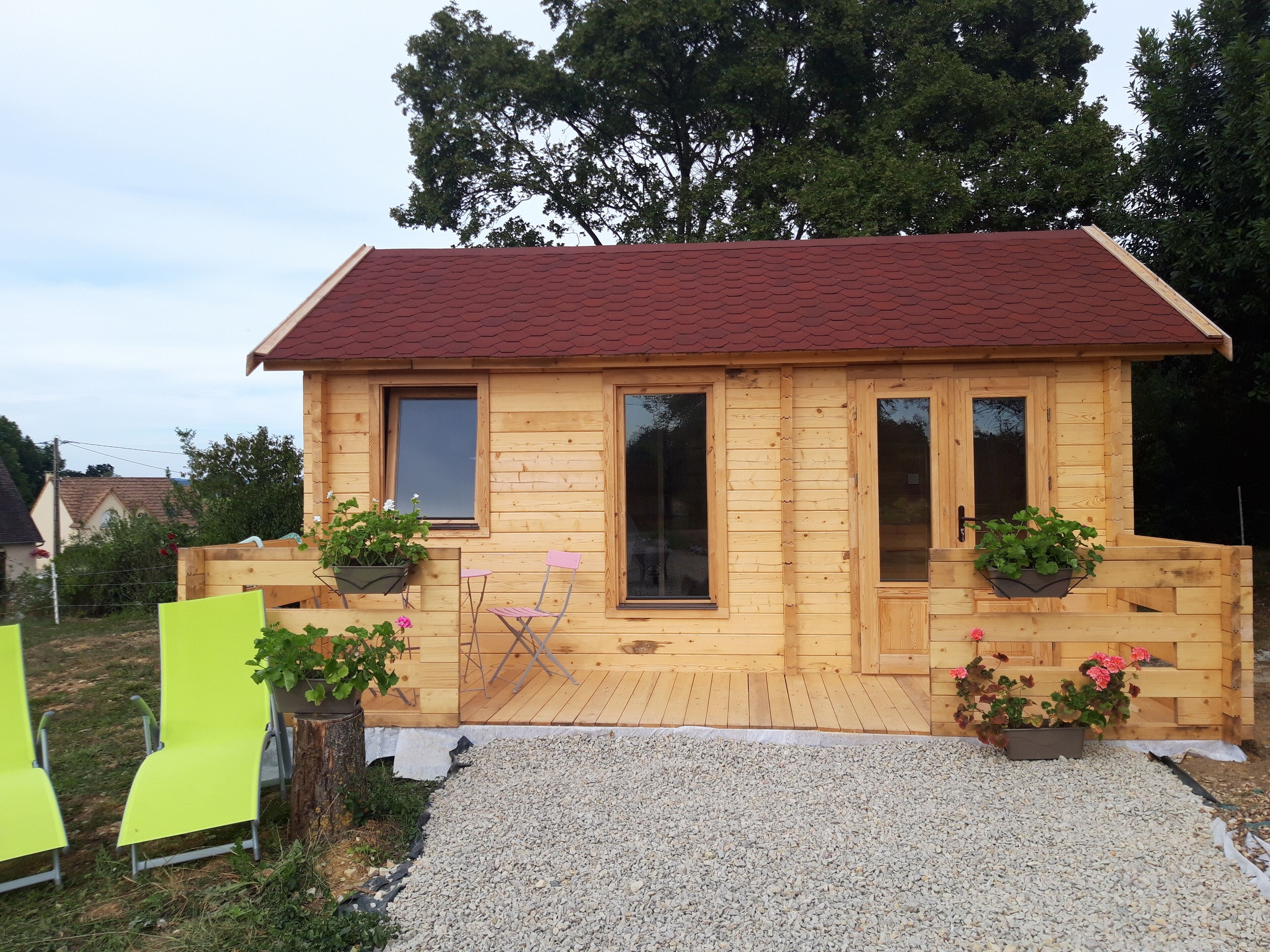 Une cabane en bois avec un toit rouge, deux fenêtres et une porte. Il y a deux plantes en pot sur le porche.