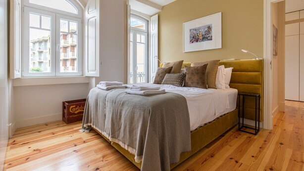 A bedroom with a large bed, a window with a view of buildings, a framed picture on the wall, and a bedside table. - Olá Lisboa Apartment (Lisboa)