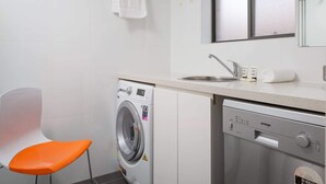 A compact laundry area with a washing machine, a dishwashing machine, a sink, and a chair. - Cosy and Bright Studio Next to Westmead Hospital (Westmead)
