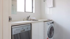 Une salle de bain moderne et compacte, dotée d’un lave-vaisselle encastré, d’une machine à laver et d’une fenêtre laissant entrer la lumière naturelle.