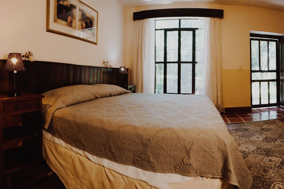 Un dormitorio con una cama grande, una mesita de noche, una lámpara, un cuadro en la pared y una ventana con cortinas.