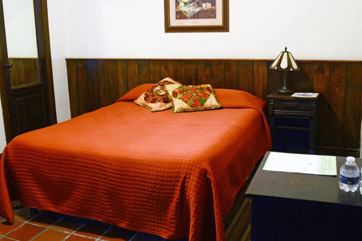 Una cama bien hecha con colcha roja, dos almohadas, una mesita de noche con lámpara y una botella de agua en el buró.
