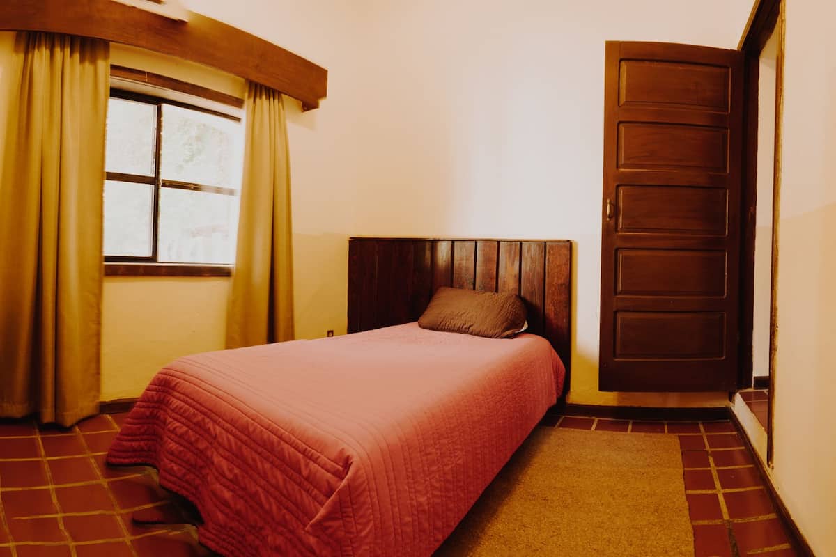 Un dormitorio con cama, cabecera de madera, mesita de noche, escritorio, ventana con cortinas y puerta.
