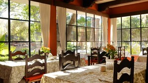 Uma área de refeições com mesas preparadas para quatro pessoas, grandes janelas com cortinas e vista para o verde lá fora.