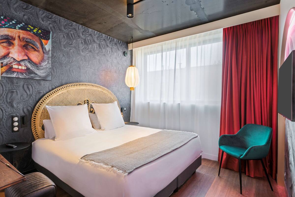 Een moderne hotelkamer met een groot bed, een rieten hoofdbord, een rood gordijn en een teal-kleurige stoel.