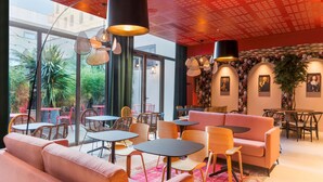 Un café moderno con un sofá rosa, sillas y mesas de madera. Se observan ventanas amplias y un techo rojo.
