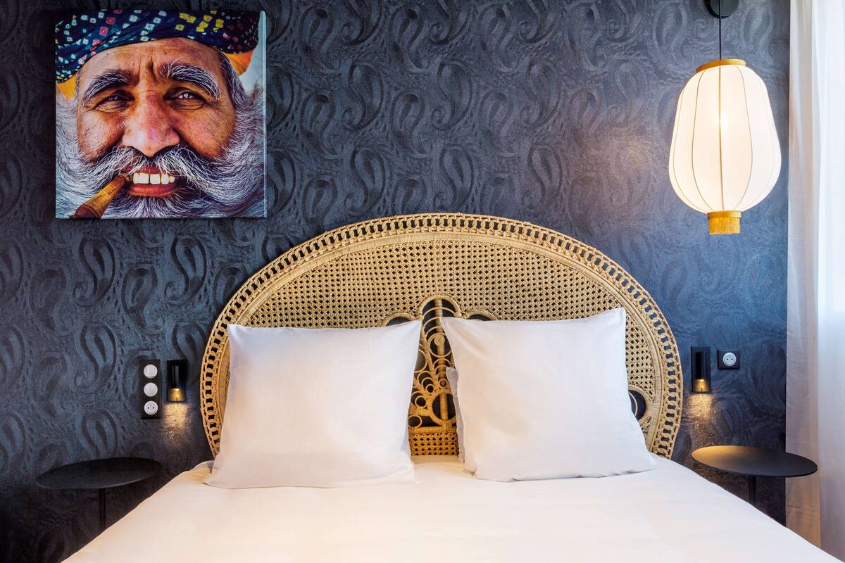 Een bed met een geweven hoofdbord, twee witte kussens, een wandlamp en een klein rond tafeltje naast het bed.