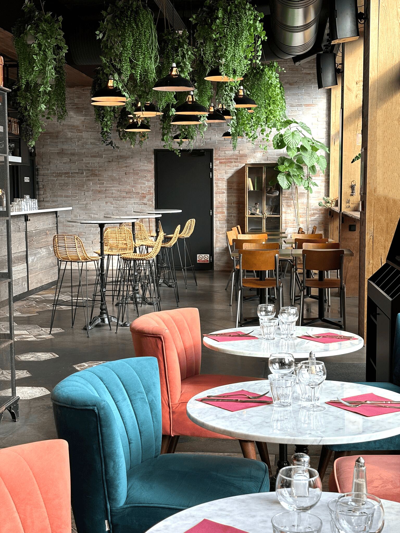 A modern restaurant interior with brick walls, hanging plants, and a variety of seating options including round tables and cushioned chairs.