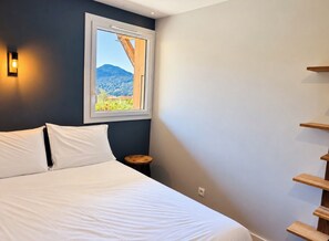 A bedroom with a bed, a window with a view of a mountain, a small round table, and a wooden shelf unit.