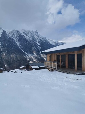A wooden cabin with a balcony, surrounded by snow-covered mountains and a clear sky.