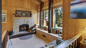 Interior acogedor de una cabaña de troncos con chimenea de piedra, vigas de madera y un amplio ventanal con vistas a un bosque.