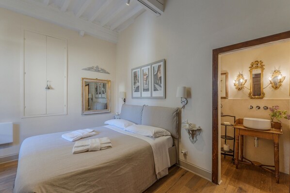 A bedroom with a large bed, a bedside table with a lamp, and a bathroom with a sink and mirror.