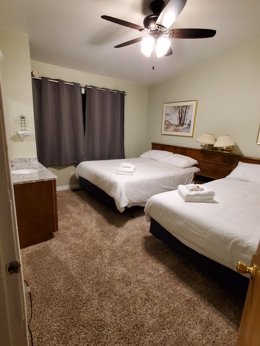 A hotel room with two beds, a ceiling fan, a painting on the wall, and a bathroom with a sink and mirror.