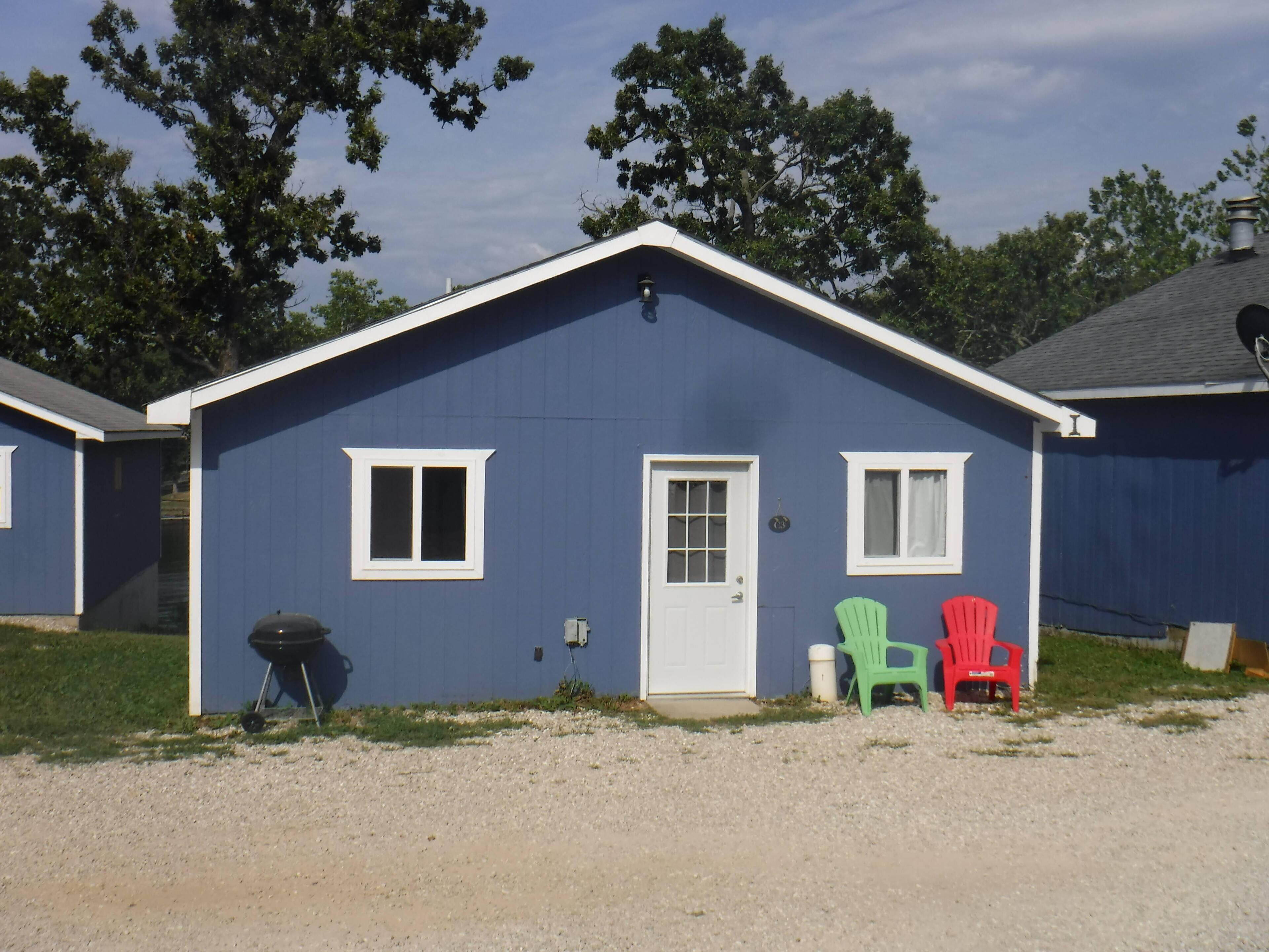 A small, single-story building with a dark blue exterior and white trim. There are two windows, one on each side of a central door. Two plastic chairs are placed outside in front of the building.