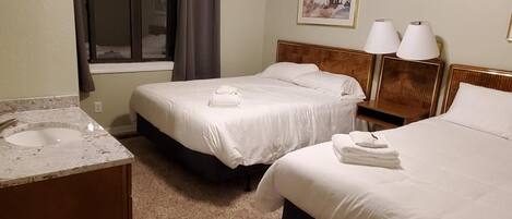 A hotel room with two beds, a ceiling fan, a window with curtains, a sink, and a bedside table with lamps.