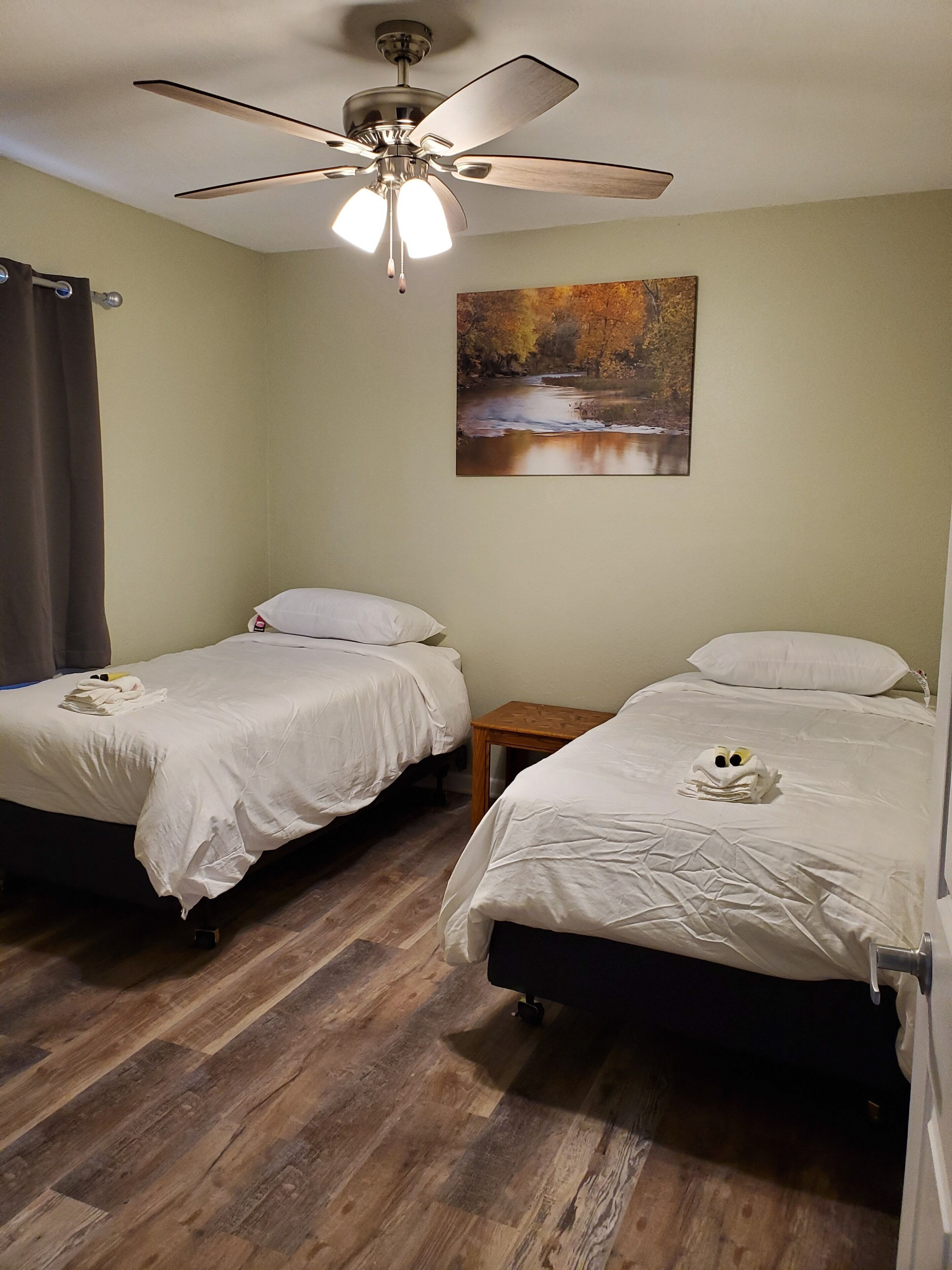 A hotel room with two beds, a ceiling fan, a painting of a river, and a wooden floor.