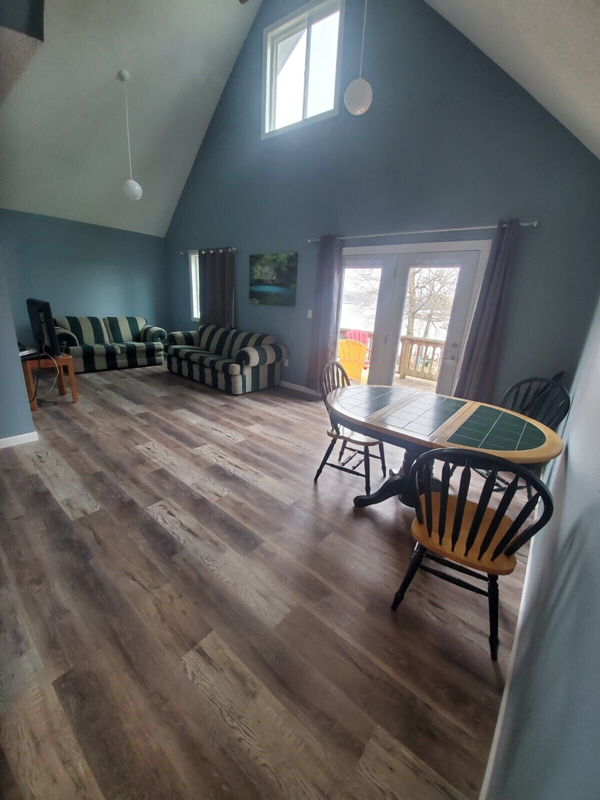 A spacious living area with a high ceiling, a dining table with chairs, a sofa set, and a view through a glass door to an outdoor deck.