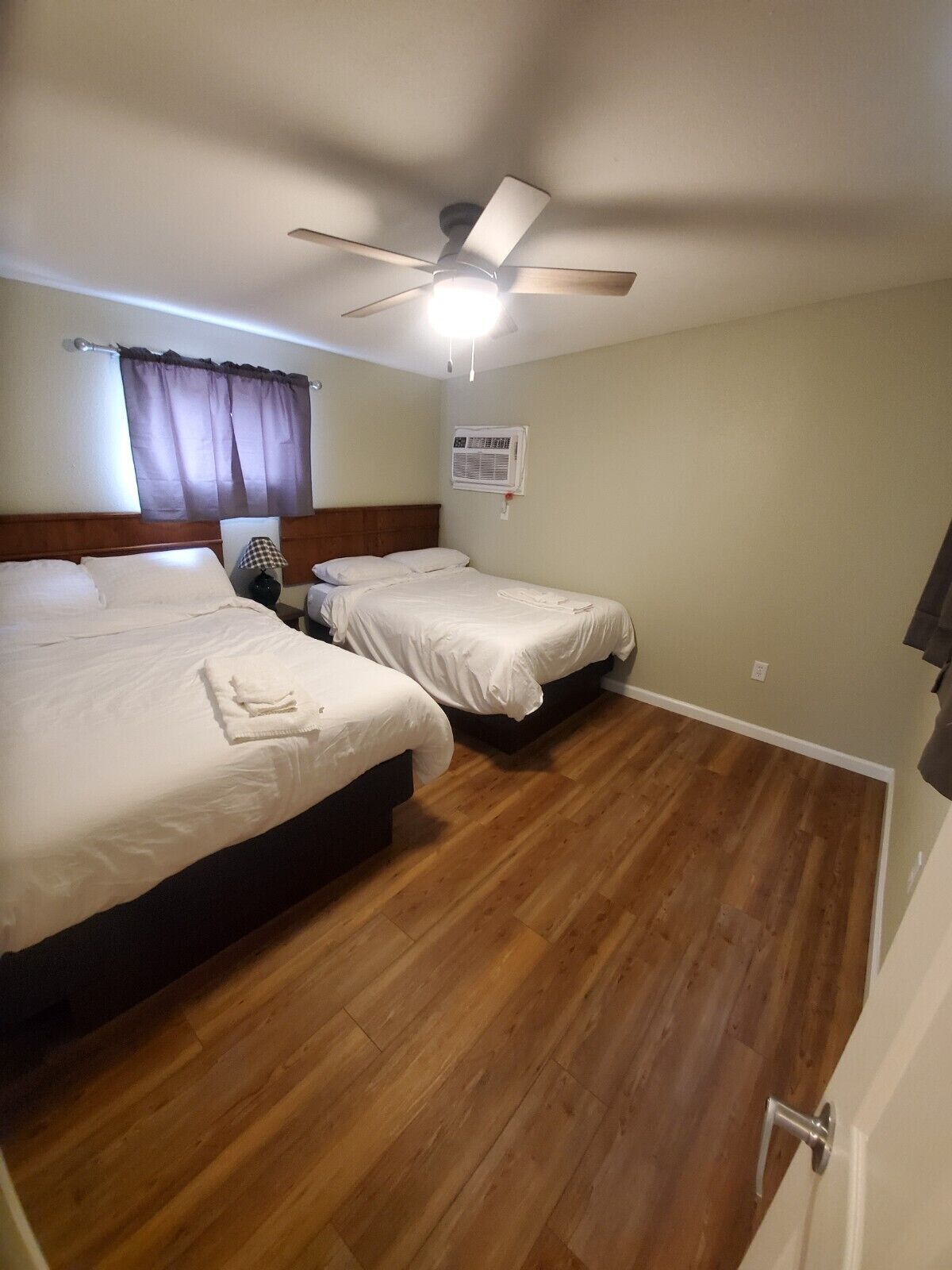 A hotel room with two beds, a ceiling fan, an air conditioning unit, and wooden flooring.