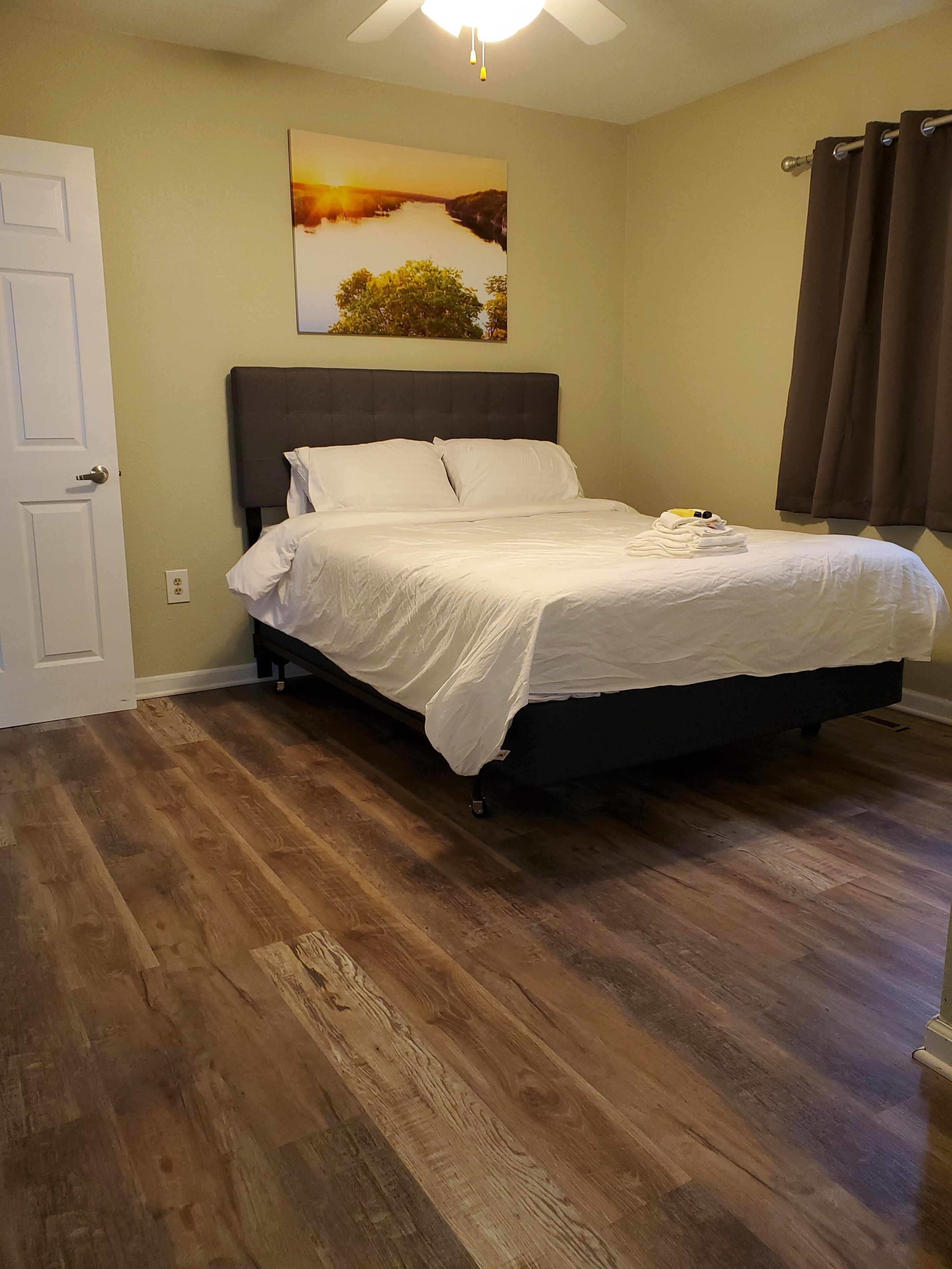 Un dormitorio con cama, piso de madera, ventilador de techo, un cuadro y una puerta.