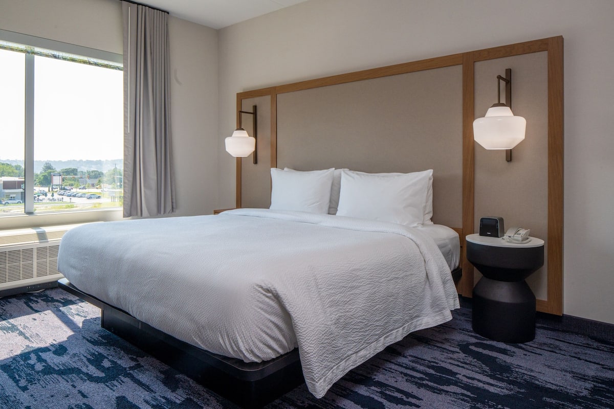 A hotel room with a large bed, two wall-mounted lamps, a bedside table with a phone, and a view of a parking lot through a window.