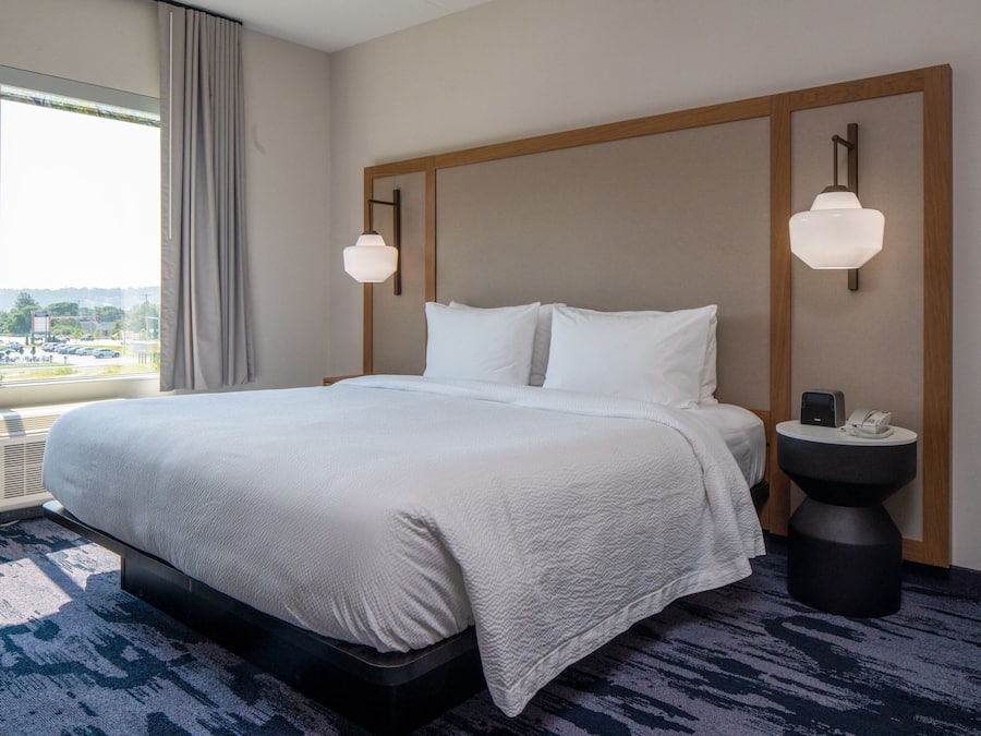 A hotel room with a large bed, two wall-mounted lamps, a bedside table with a phone, and a view of a parking lot through a window.