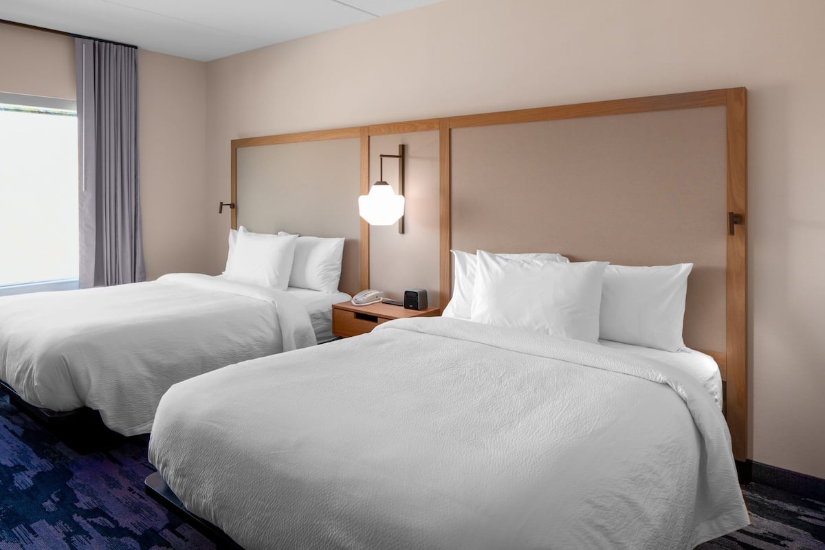 A hotel room with two beds, a nightstand with a phone and a lamp, and a large window with curtains.