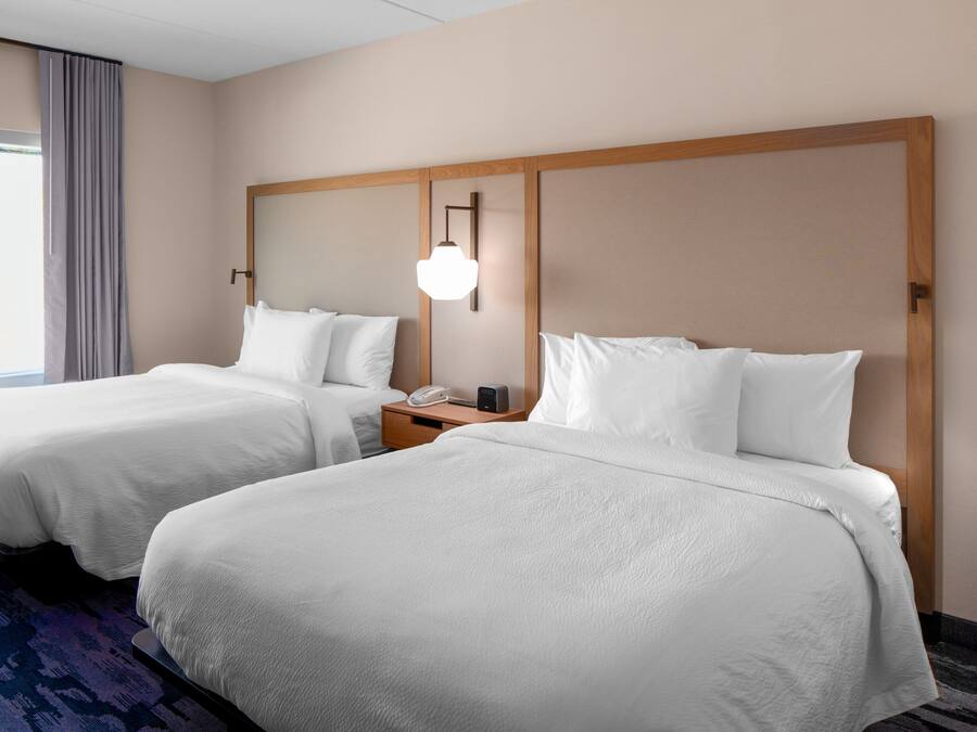 A hotel room with two beds, a nightstand with a phone and a lamp, and a large window with curtains.