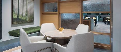 A modern meeting room with a round table, four chairs, a green bench, and a large window with a view of the dining area.
