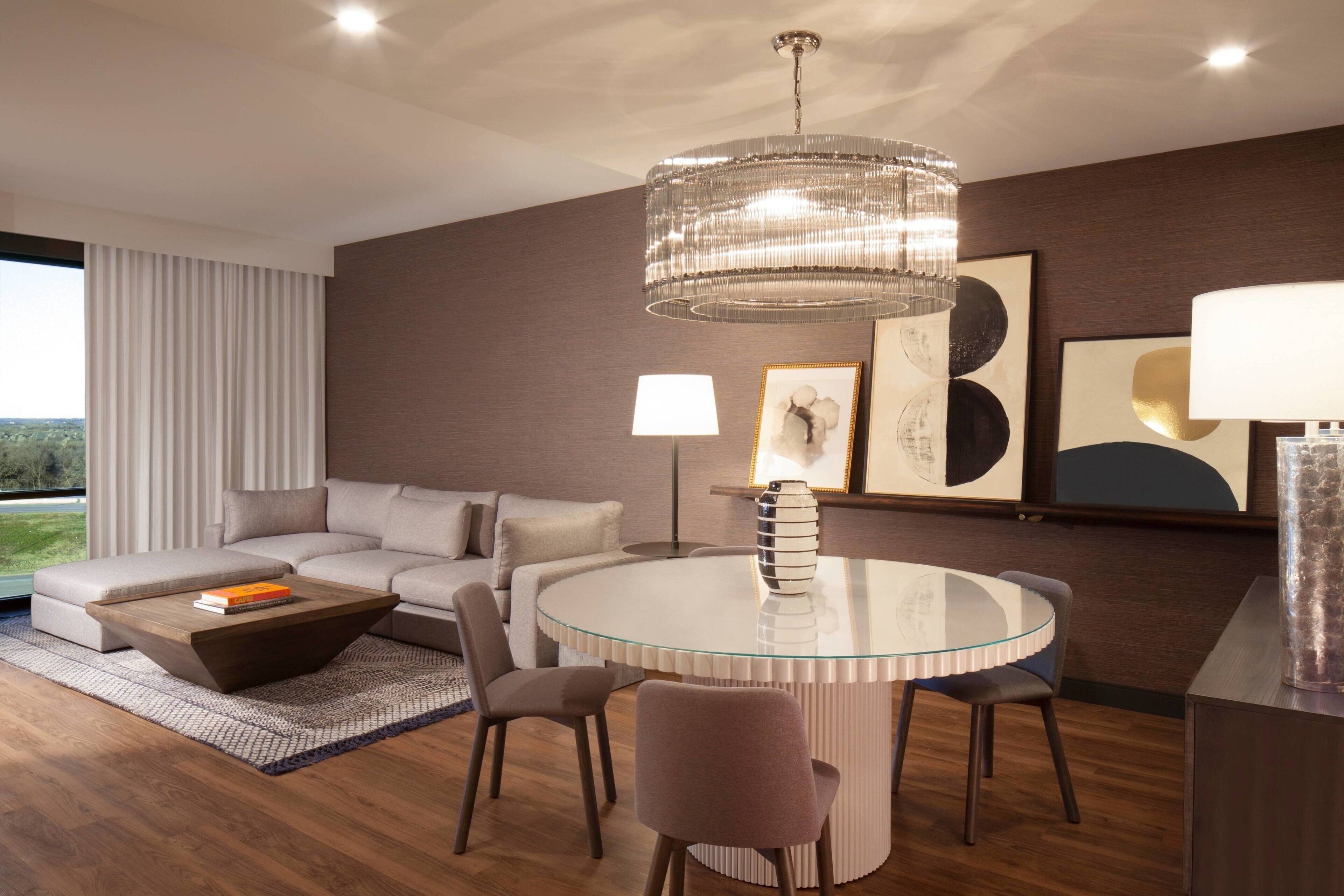 A modern living room with a glass coffee table, a round dining table, and a chandelier. There is a sofa and a lamp in the room.
