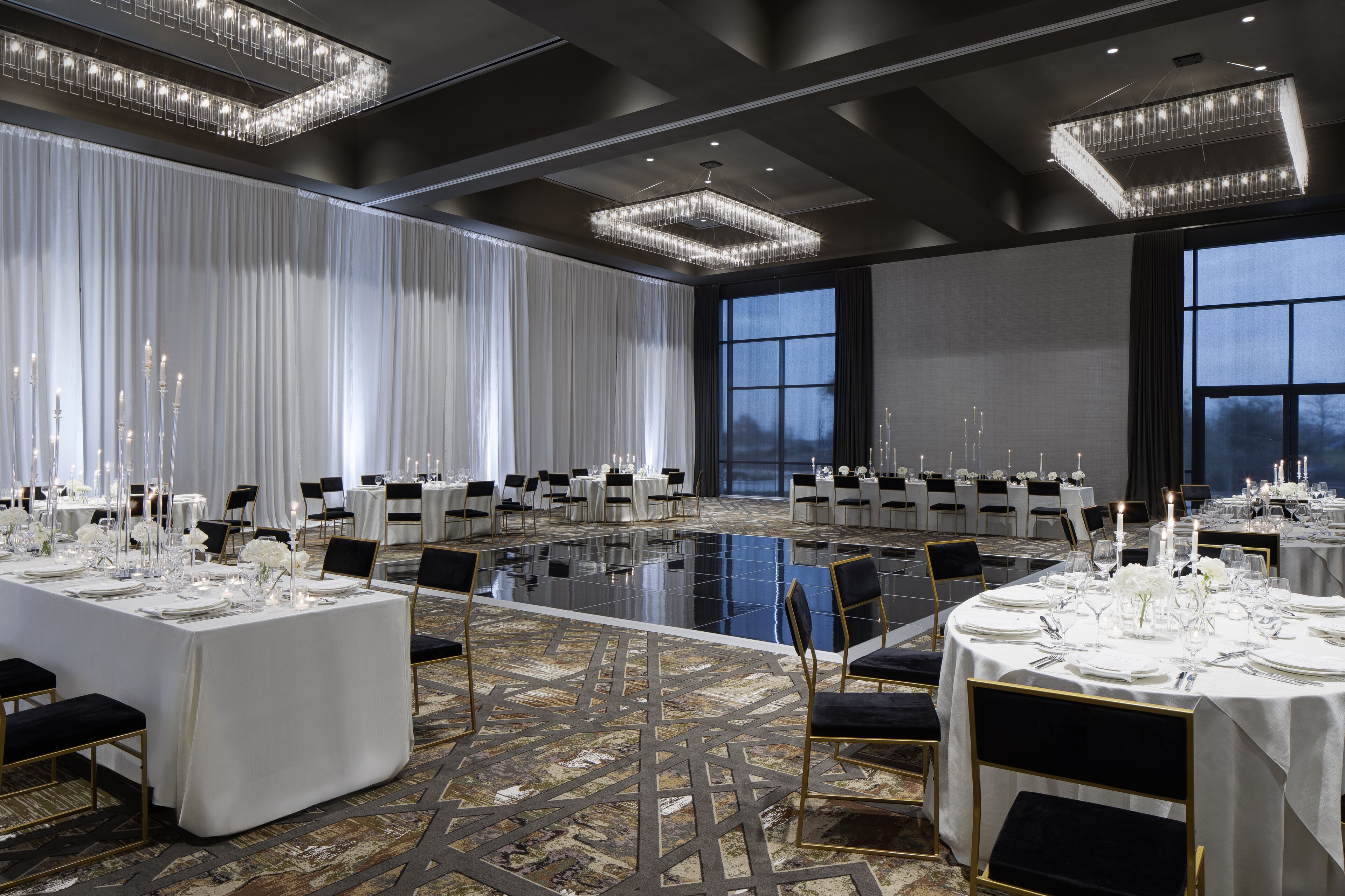 A spacious banquet hall with multiple round tables set for dining, large windows, and modern chandeliers.
