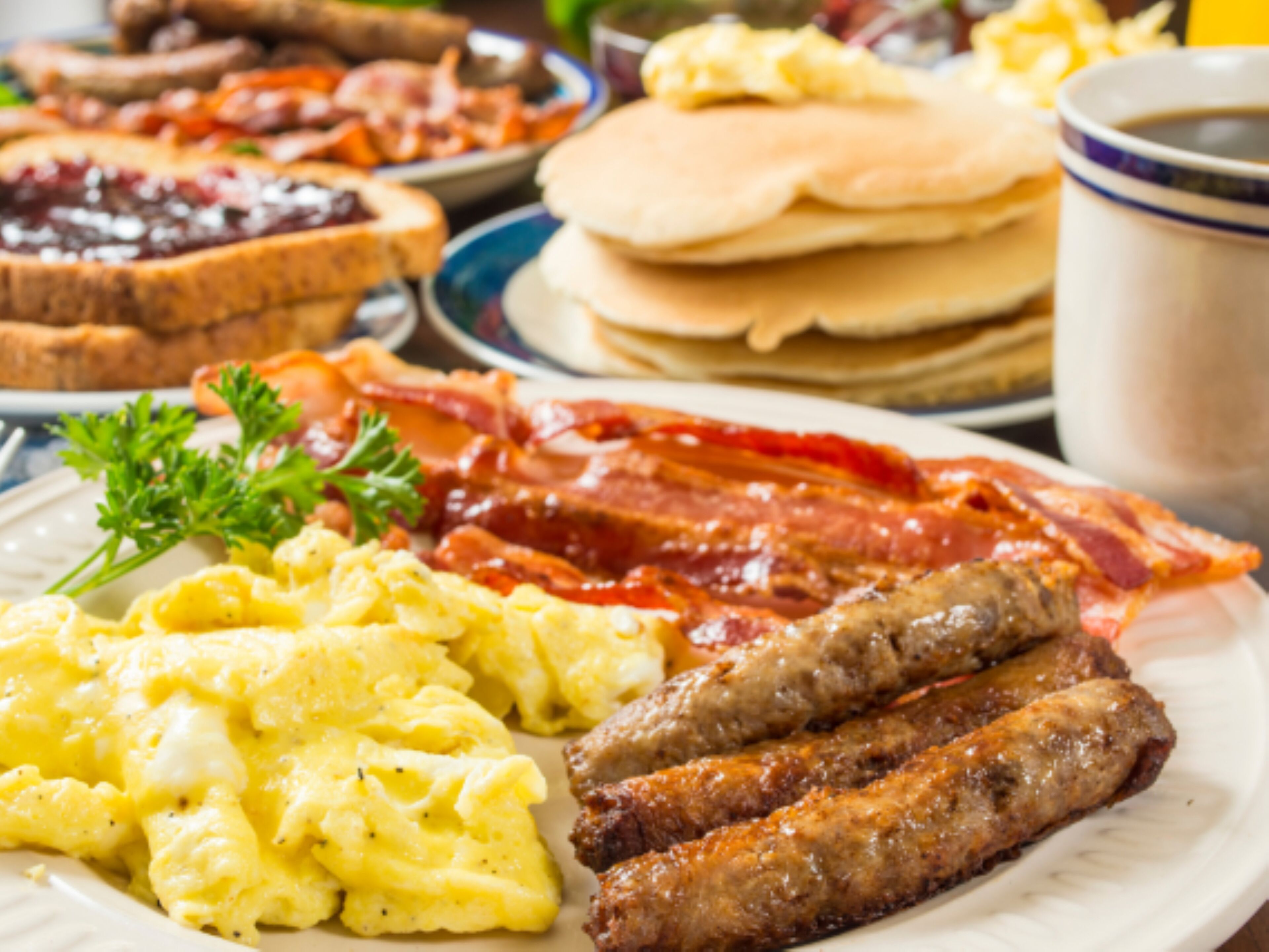 A breakfast plate with scrambled eggs, sausages, bacon, toast, and a cup of coffee.
