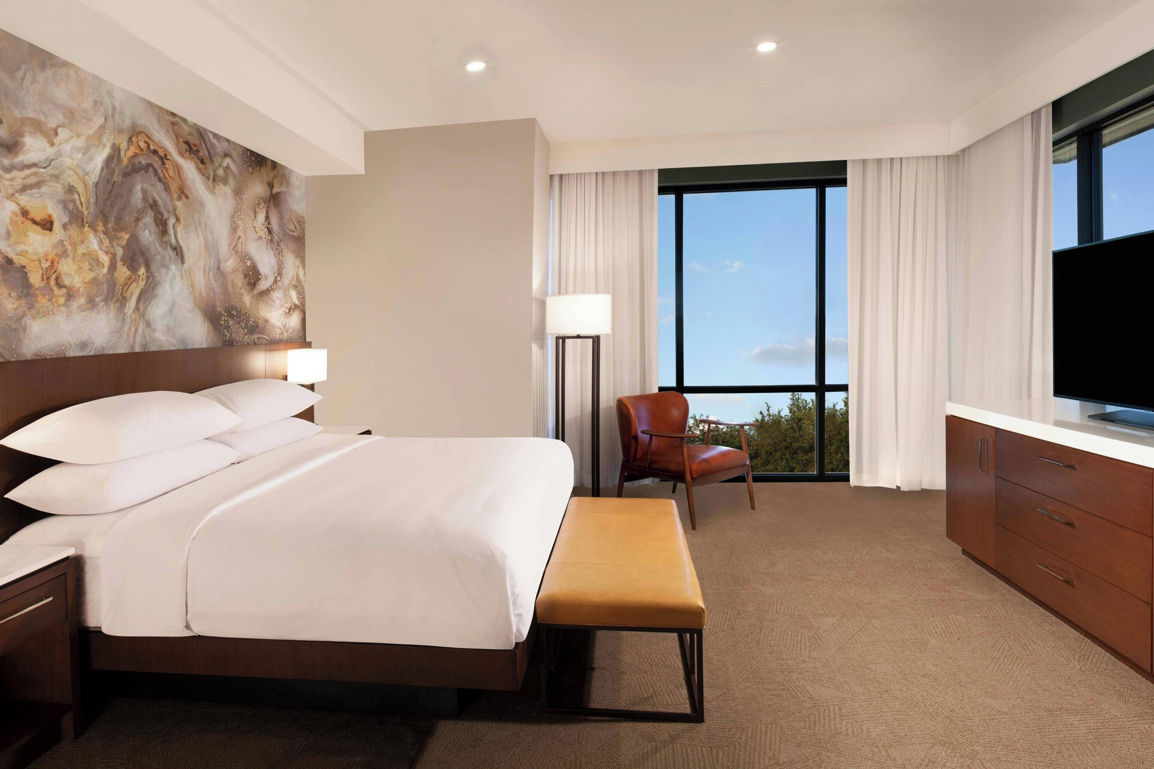 A hotel room with a large bed, a desk, a chair, a TV, and a view of the outdoors.