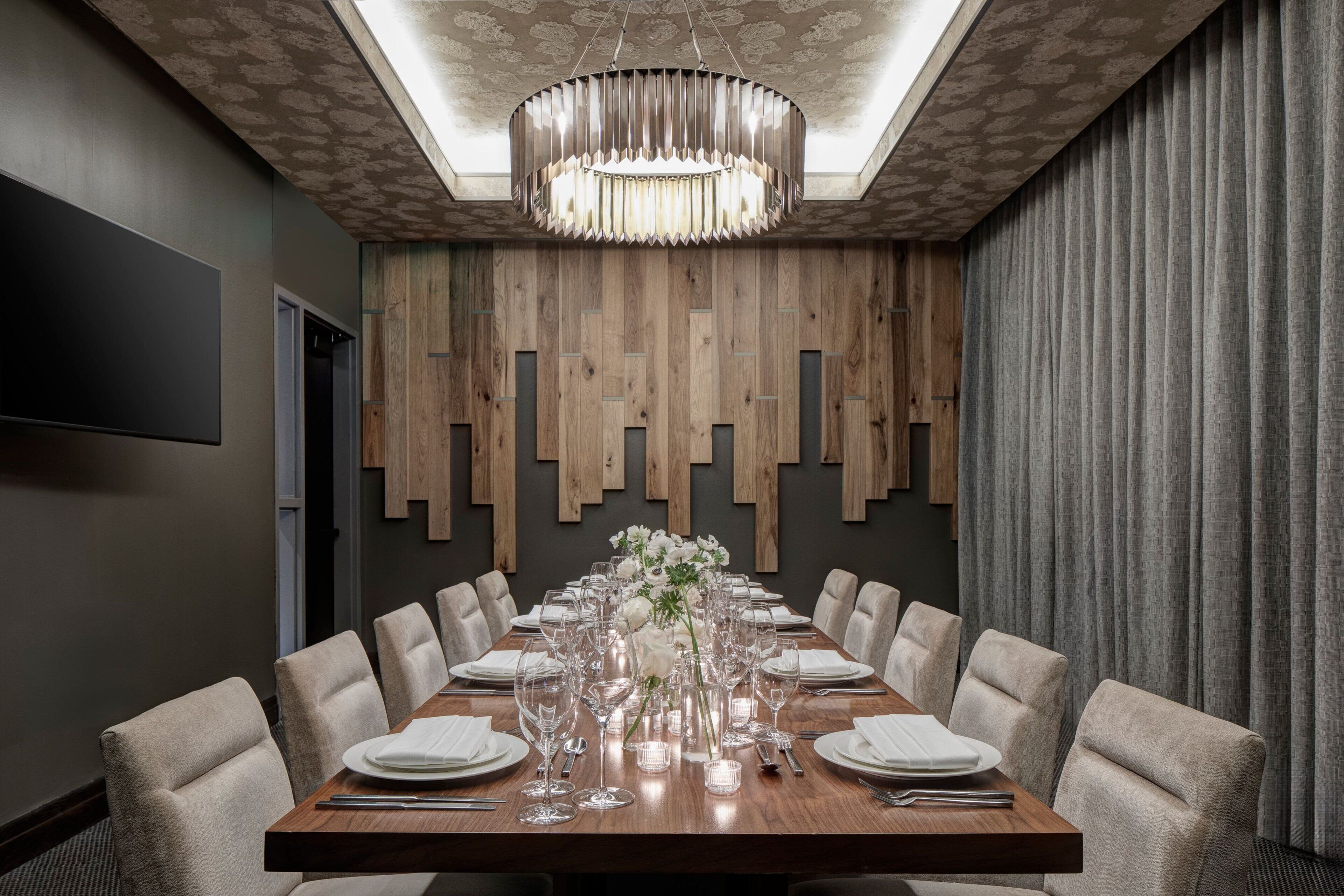A conference room with a long wooden table set for a meeting, surrounded by chairs, with a modern chandelier and a patterned ceiling.