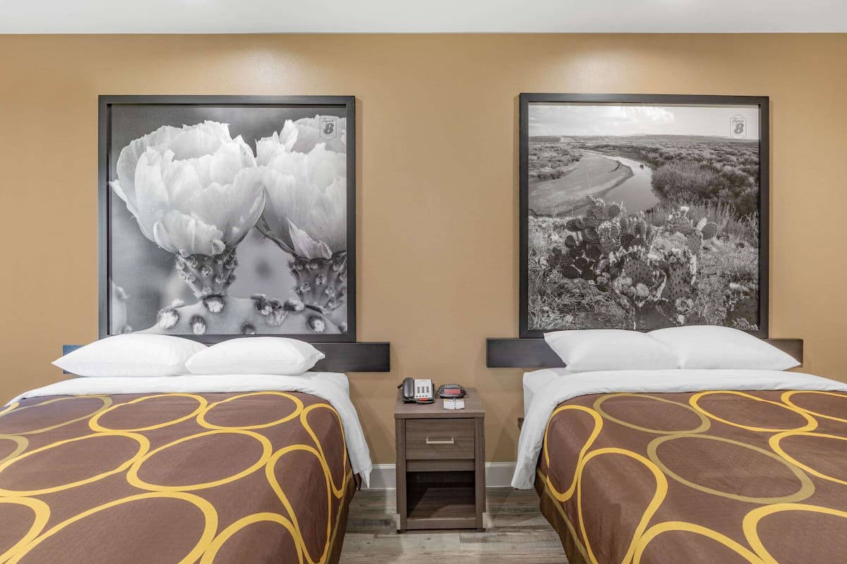 A hotel room with two beds, each with a patterned bedspread, a nightstand with a phone and a small table, and two large black and white landscape paintings on the walls.