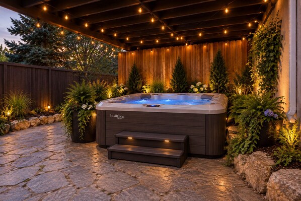 A hot tub with a wooden deck and stone pathway, surrounded by plants and a wooden fence.