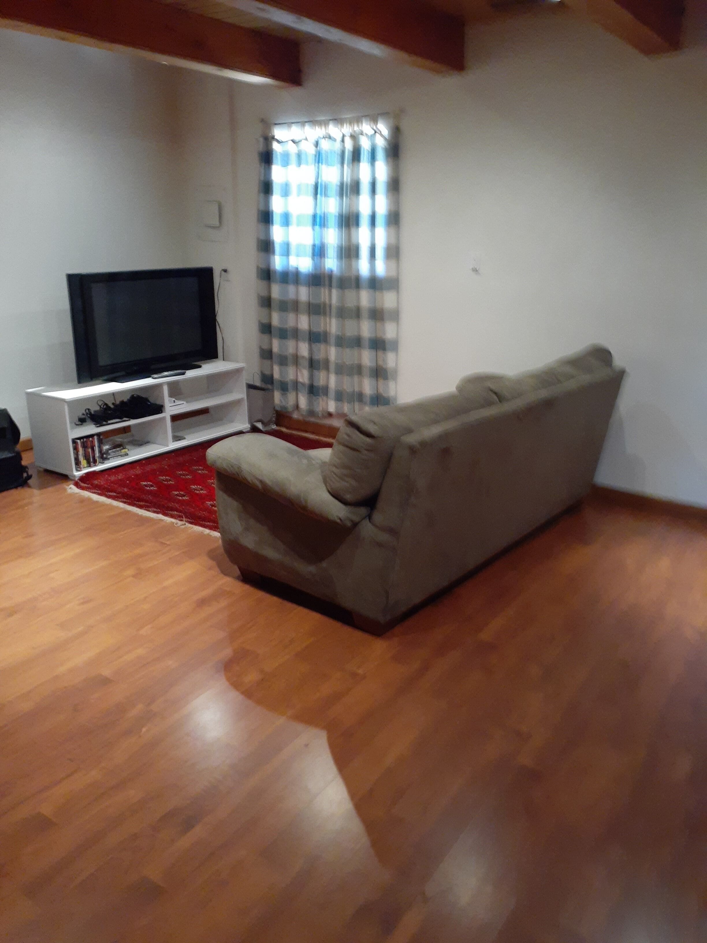 Una sala de estar con un sofá, un televisor sobre un mueble, una alfombra roja y una ventana con cortinas a cuadros.