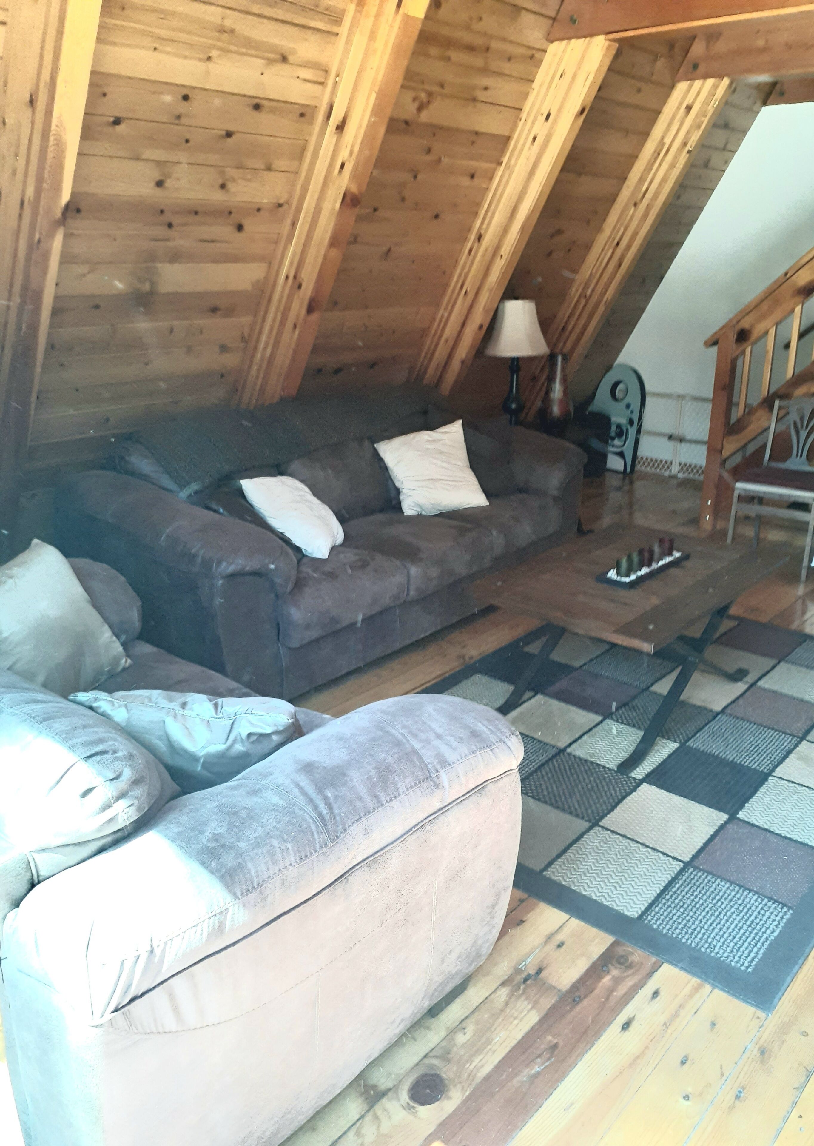 A cozy loft-style room with a wooden ceiling, a gray sofa, a matching armchair, and a patterned rug.