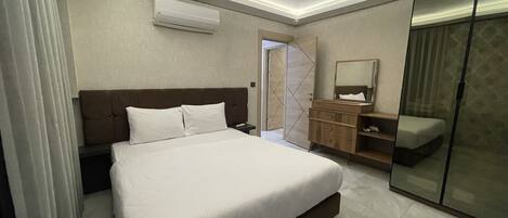 A modern hotel room with a large bed, a wardrobe, a vanity with a mirror, and an air conditioning unit.