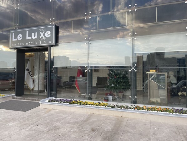Exterior del Hotel & Spa Le Luxe Suites, con fachada moderna de vidrio, entrada visible y un pequeño árbol de Navidad.