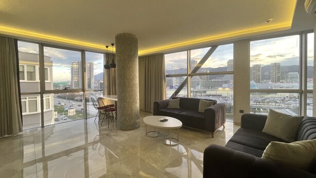 A spacious living room with a high ceiling, large windows offering a city view, a central pillar, and a modern seating arrangement. - Le Luxe suites hotel & SPA (Bursa)