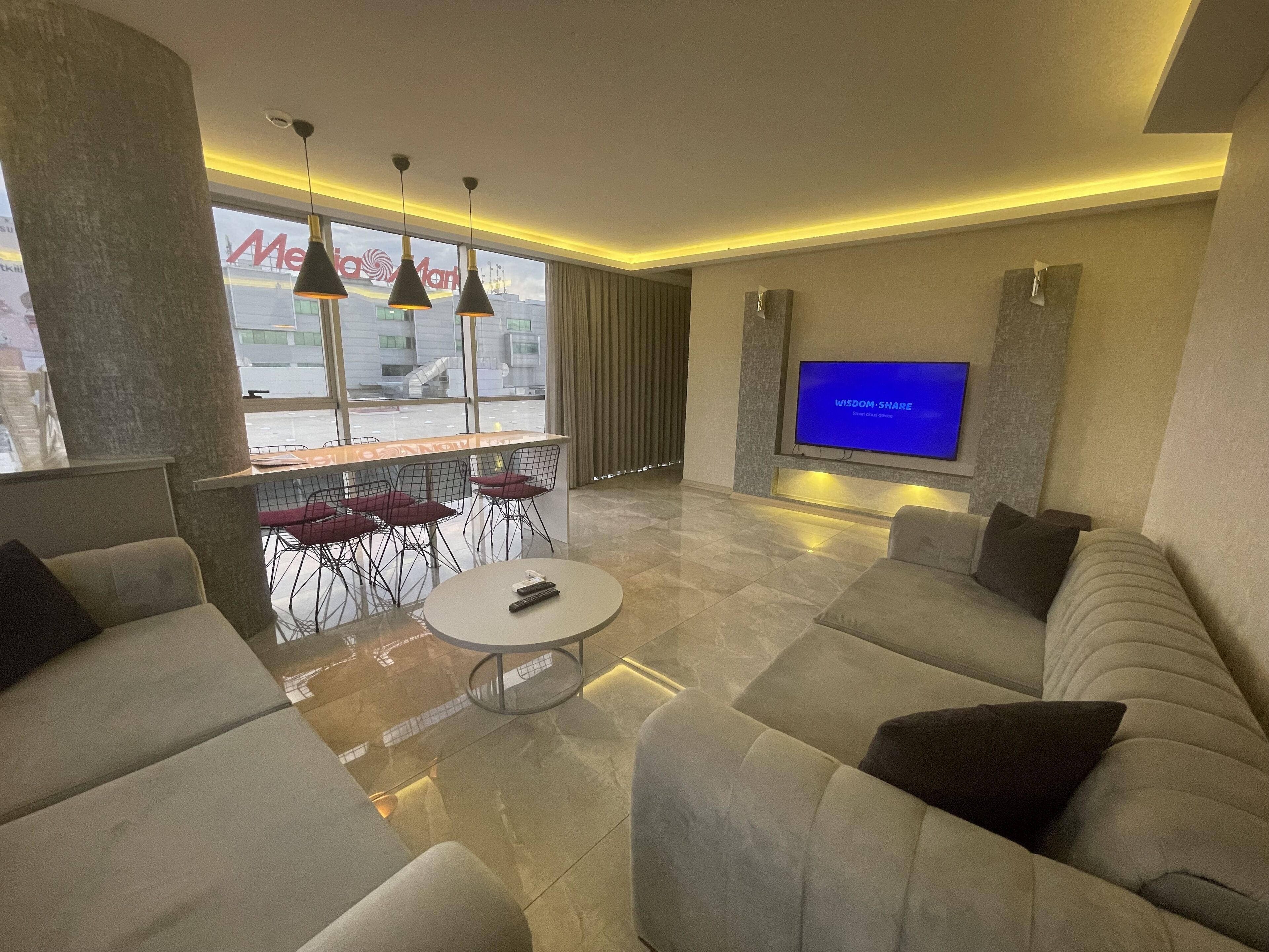 Ein modernes Wohnzimmer mit großem Flachbildfernseher, bequemen Sitzmöglichkeiten, einem Essbereich mit Stühlen und Blick ins Freie durch große Fenster.