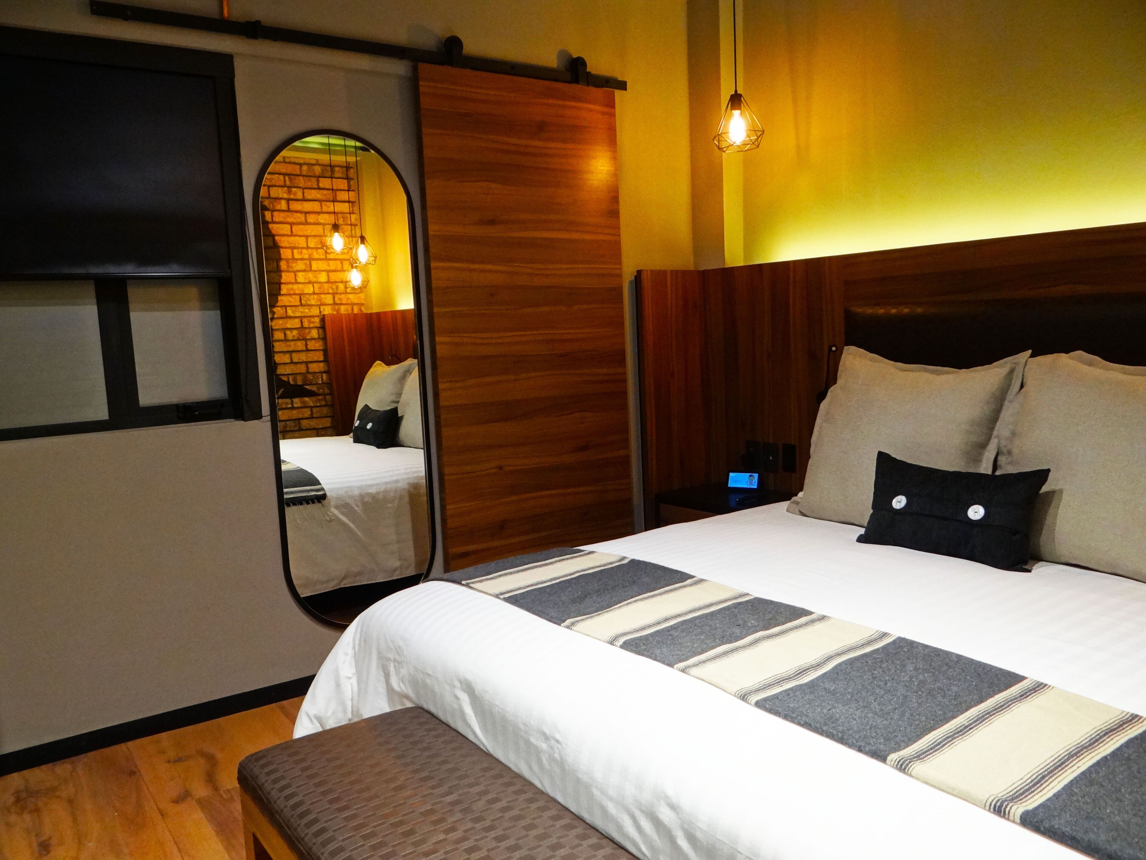 A hotel room with a bed, a bench, a nightstand, a mirror, and a wooden wall.
