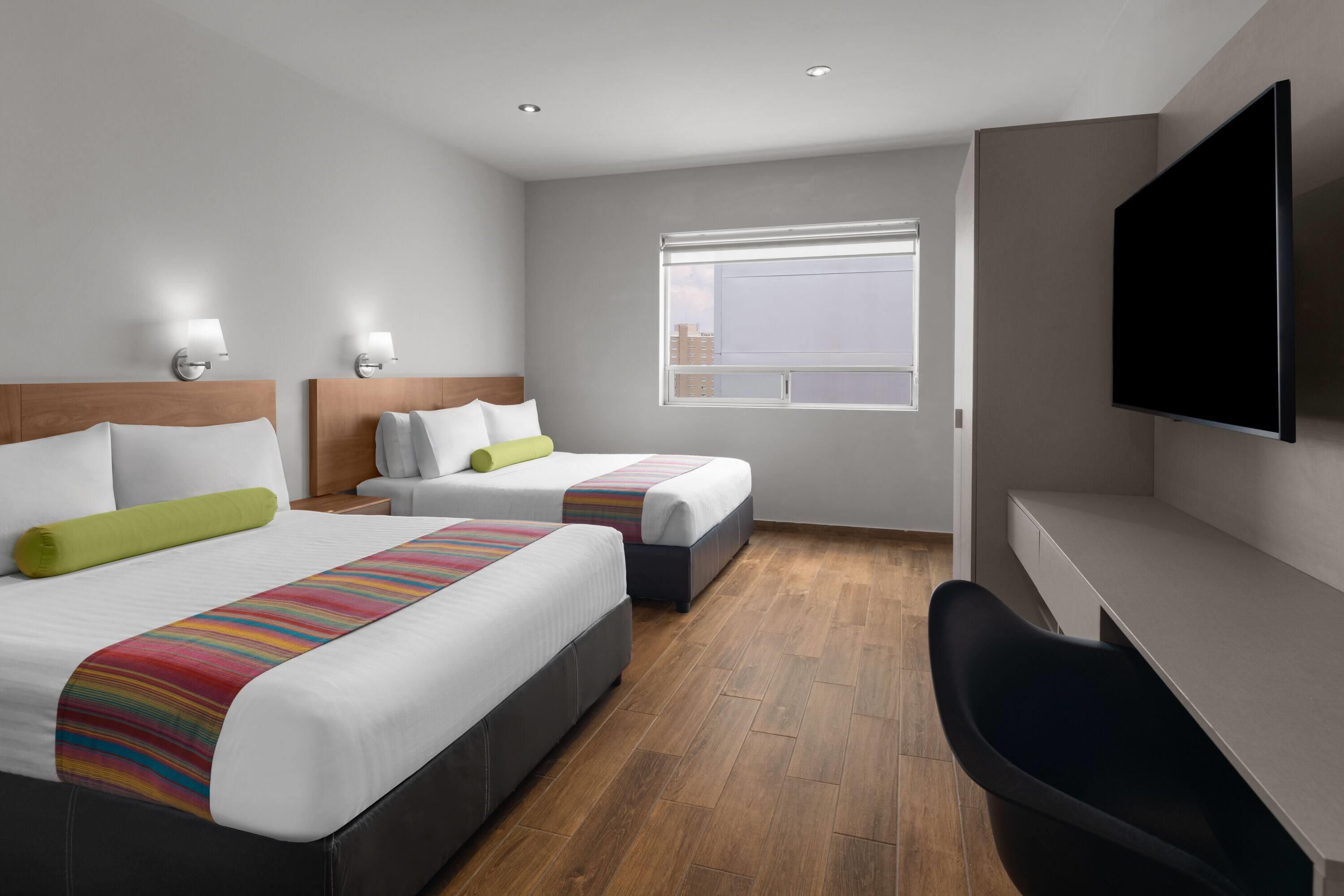 A hotel room with two beds, a flat-screen TV, a desk, and a window with a city view.